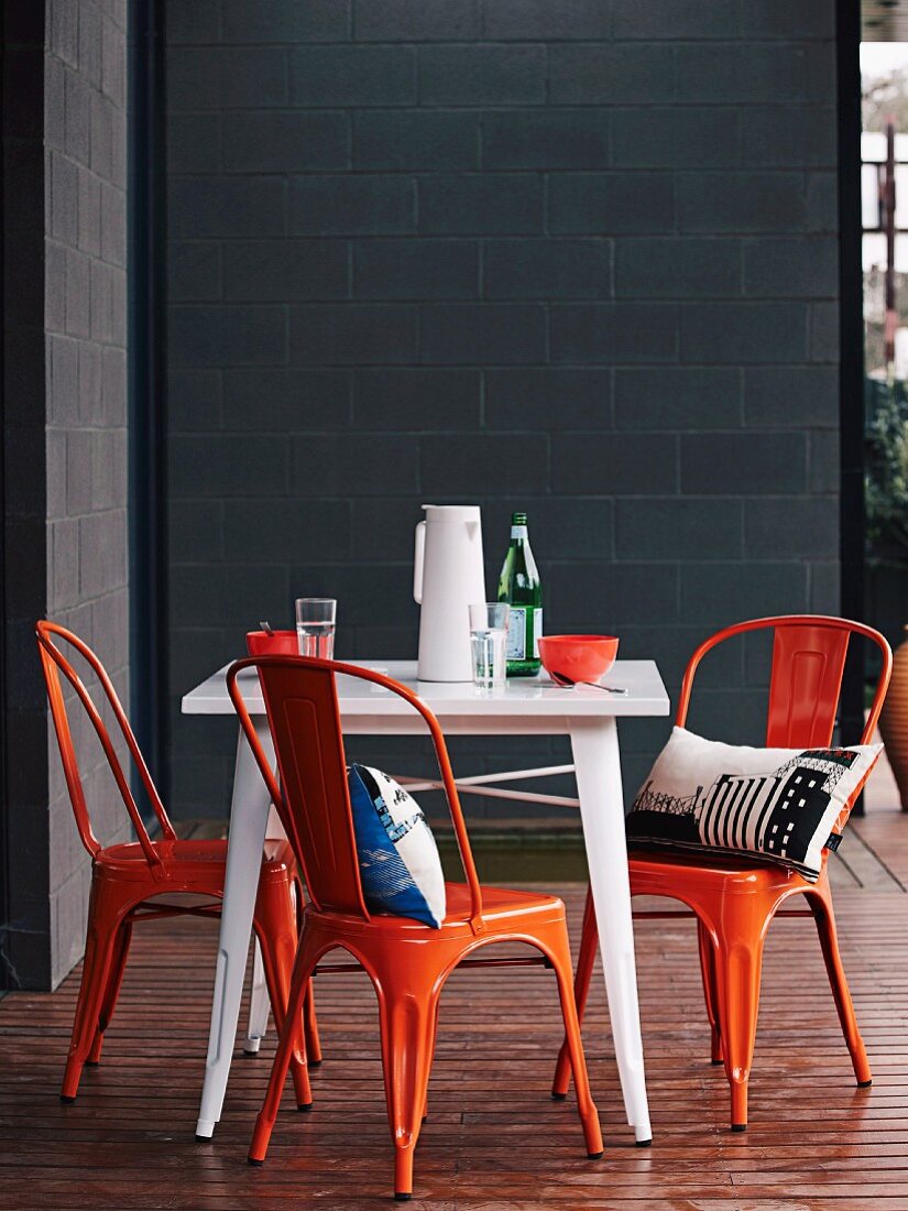 Orangefarbene Vintagestühle und weisser Tisch auf Holzterrasse vor grauer Betonsteinfassade eines Architektenhauses