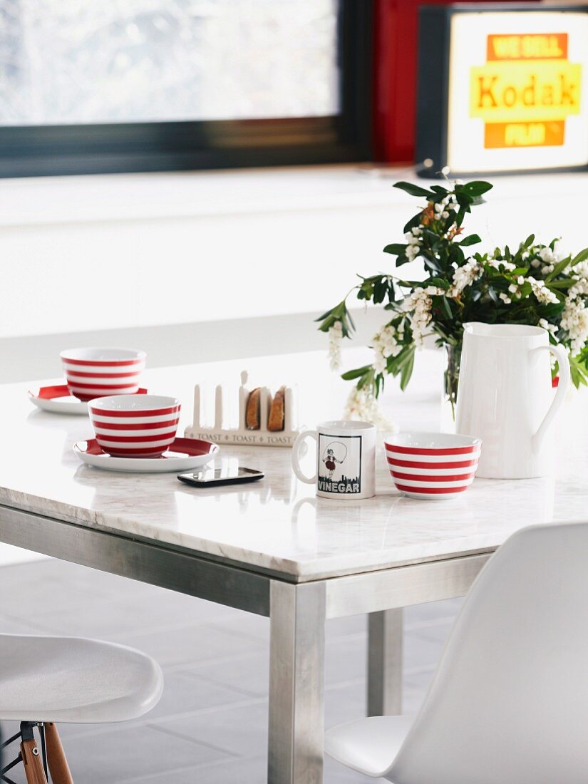 Rot-weiss gestreifte Tassen auf Esstisch mit Marmorplatte und Metallgestell, dazu klassische Schalenstühle von Eames