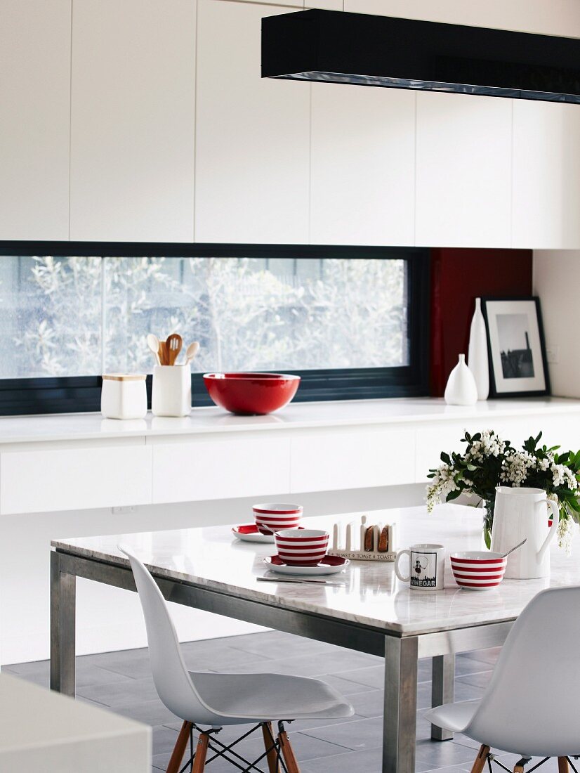 Rot-weiss gestreifte Tassen auf Esstisch mit Marmorplatte und Metallgestell, dazu klassische Schalenstühle von Eames; im Hintergrund Einbauküche