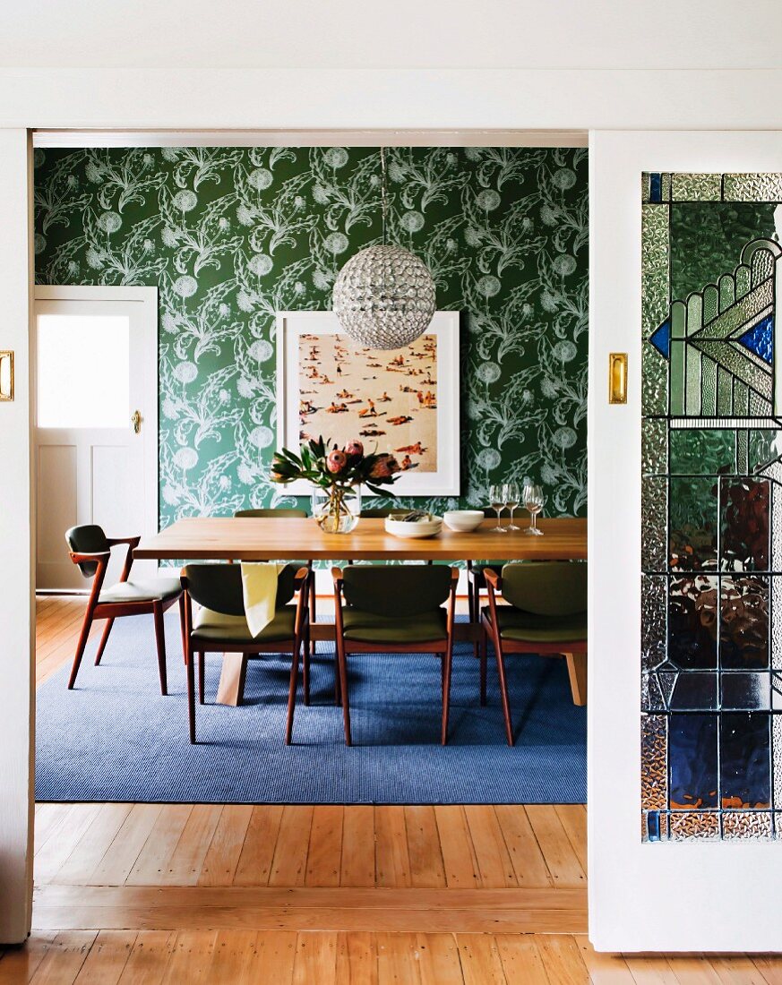 View through open sliding door into dining room with upholstered chairs around table and green wallpaper with white floral pattern