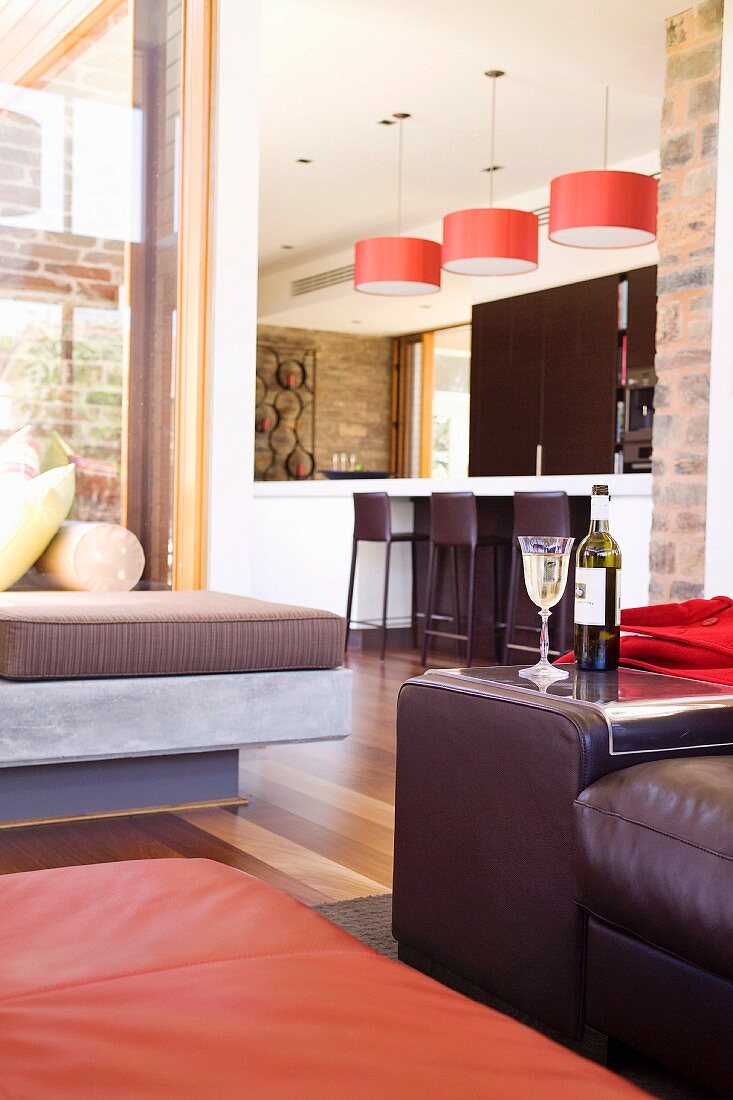 Ausschnitt eines Loungebereichs mit lederbezogenen Sitzmöbeln, im Hintergrund offene Küche mit thekenartigem Esstisch unter roten Hängeleuchten