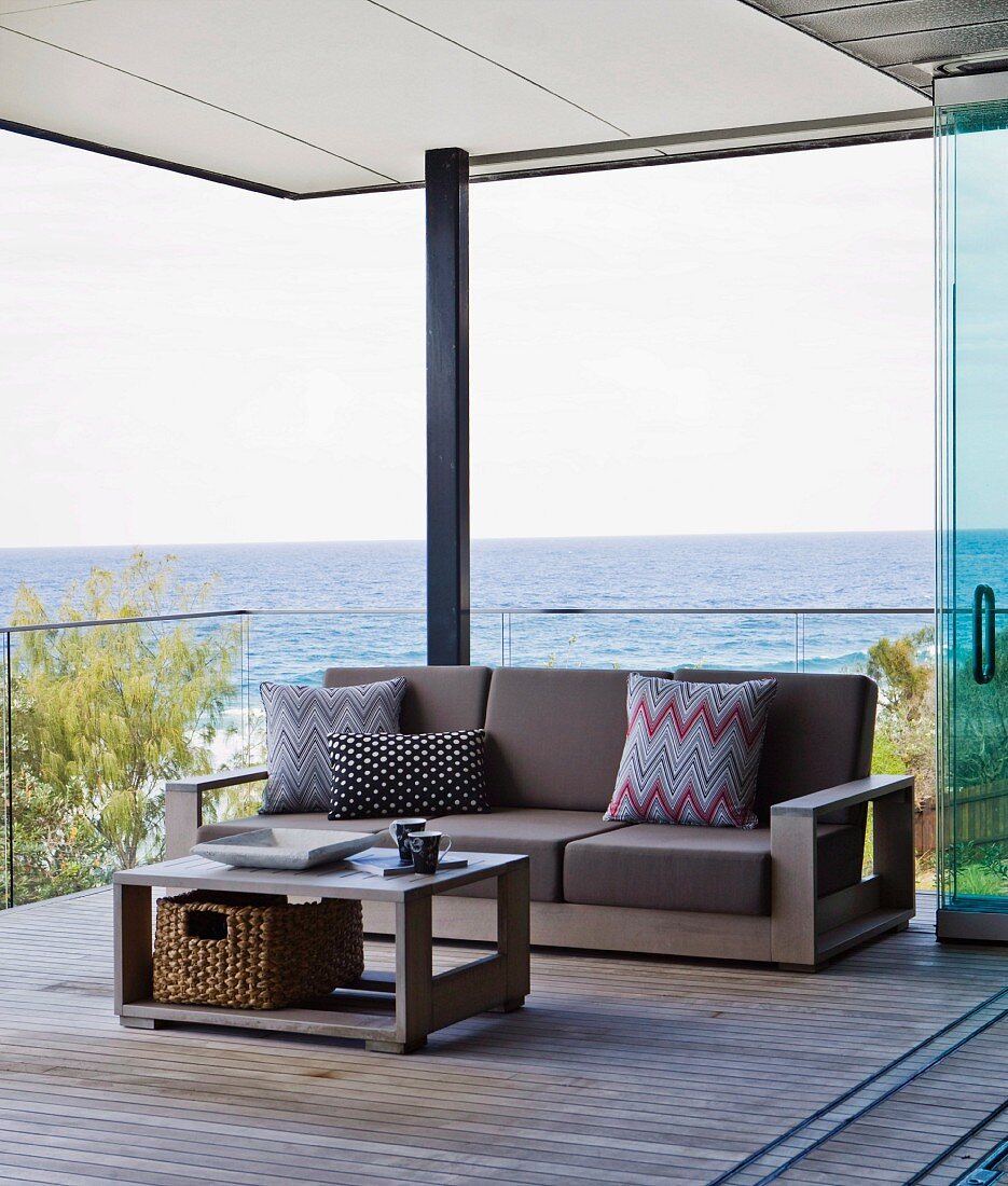 Coffee table and matching sofa with brown cushions and patterned scatter cushions against glass balustrade in relaxation area on roofed wooden deck with view of Pacific Ocean