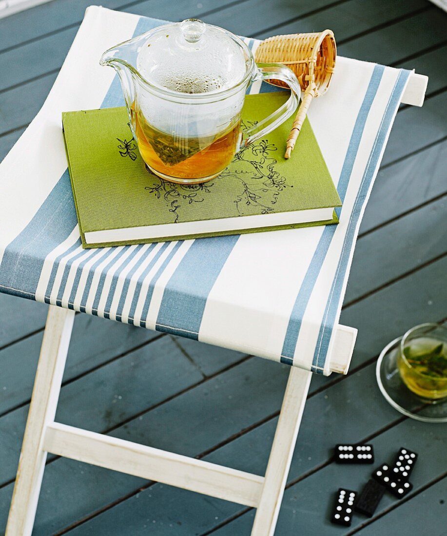 Glas Teekanne und Buch auf Hocker mit weissblau gestreiftem Bezug auf grauem Dielenboden