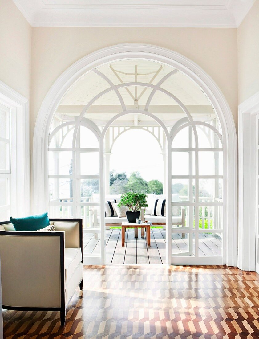 Blick auf Terrasse durch grosses Glaselement mit Rundbogengestaltung, davor ein eleganter Sessel im 20er Jahre Stil und Intarsienparkett