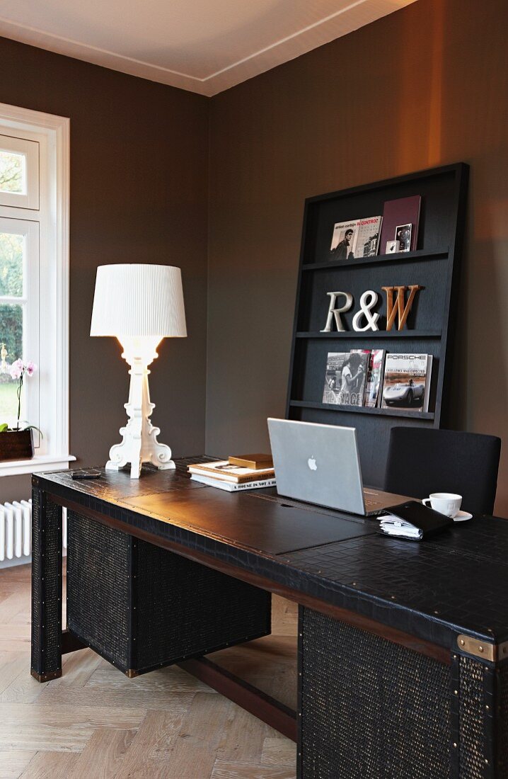 Laptop und postmoderne Leuchte auf lederbezogenem Schreibtisch; im Hintergrund Zeitschriftenregal vor warmgrau getönter Wand