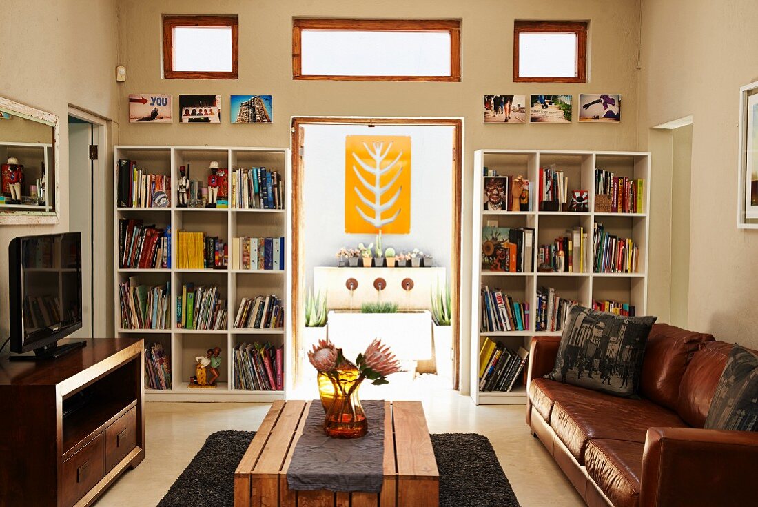 Wohnzimmer mit Ledercouch und Holztisch; Blick durch offene Terrassentür mit flankierenden Regalen auf ein dekoratives, gelbes Wandobjekt