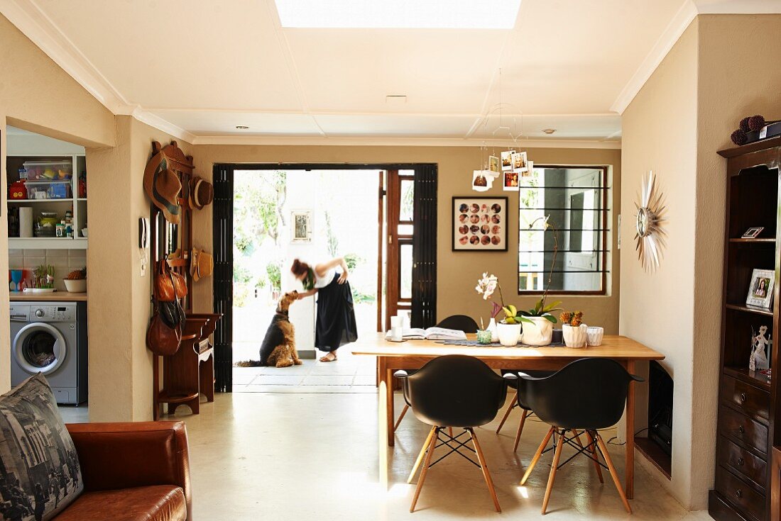 Essplatz mit Klassikerstühlen und Wohnbereich mit Ledersofa im offenen Grundriss eines Hauses; Garderobe neben weit geöffneter Haustür mit Frau und Hund im Hintergrund