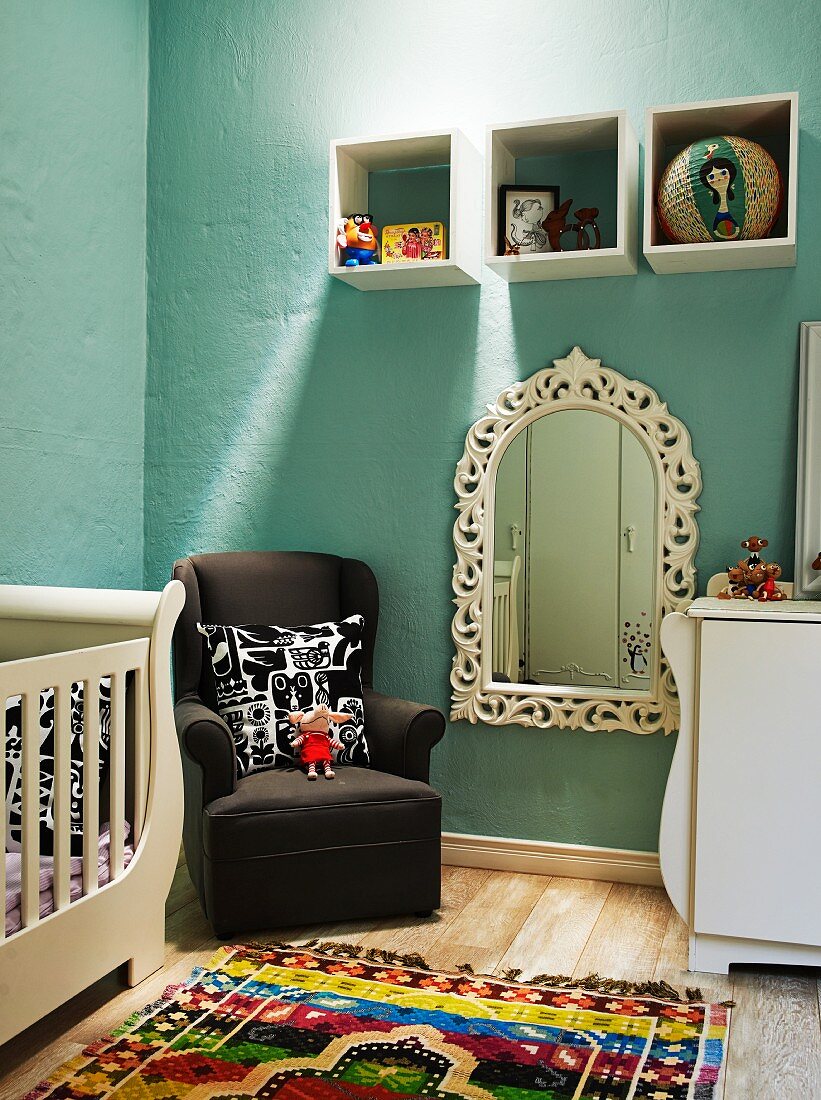 Elegantes Kleinkinderbett mit Wickelkommode in pastellblau getöntem Kinderzimmer; dekorierte Wandkästen über Stillsessel und romantischem Wandspiegel