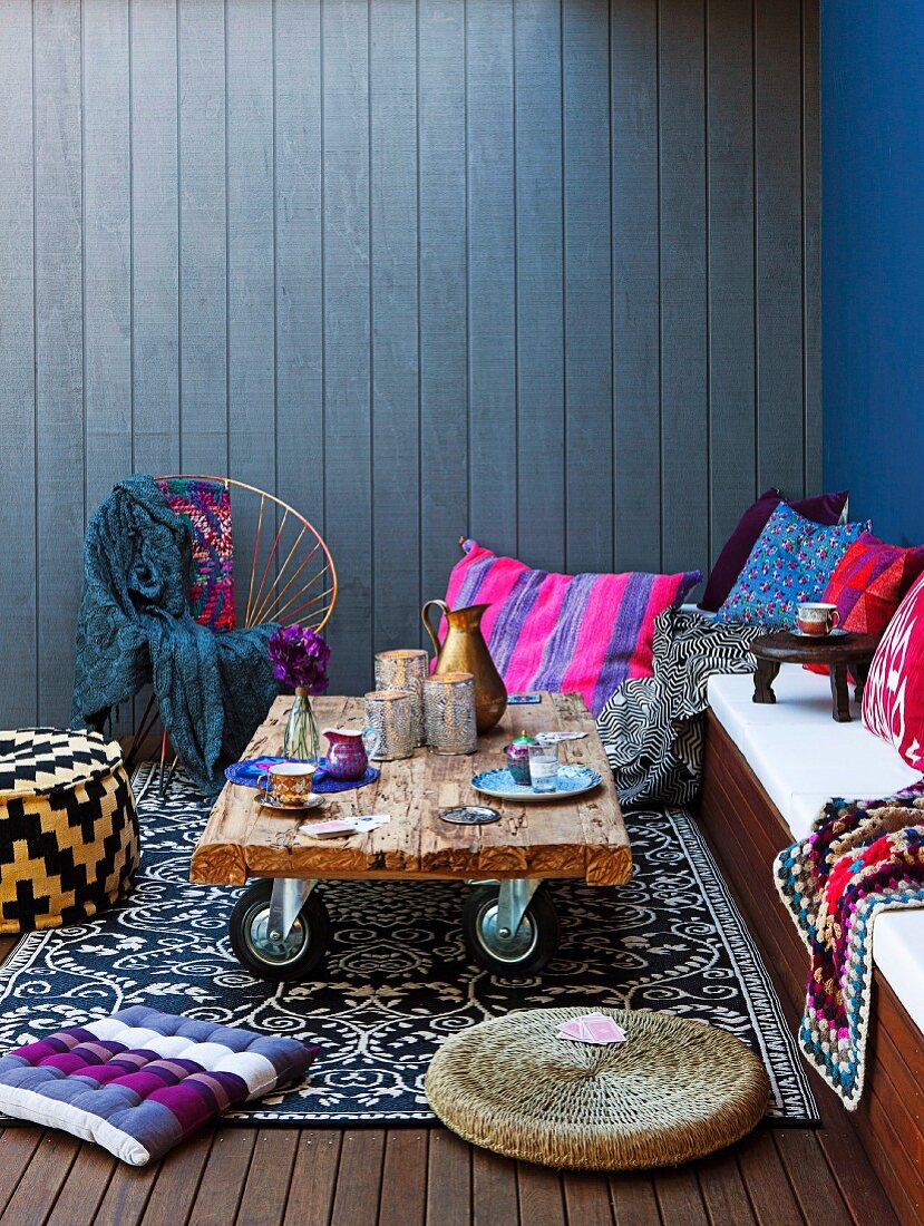 Upholstered bench with colourful scatter cushions and rustic side table on castors against blue, wood cladding