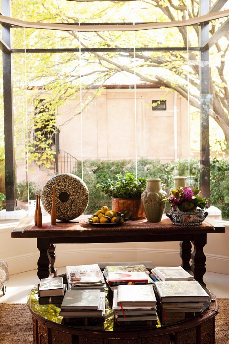Bücherstapel auf Bodentisch vor antikem Holztisch mit Blumen und Vasen in kreisförmigem Erker und Blick in Innenhof