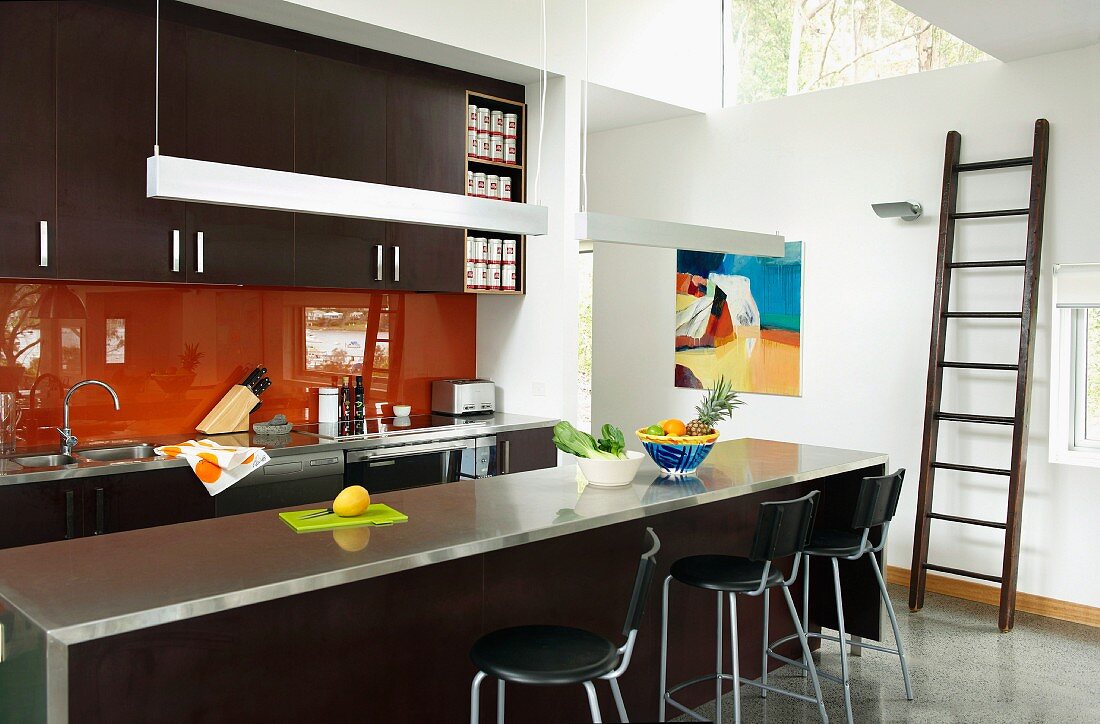 Dunkelbraune Designerküche mit orangefarbenem Glas-Spritzschutz, Edelstahl Arbeitsplatte auf Theke mit Barhockern