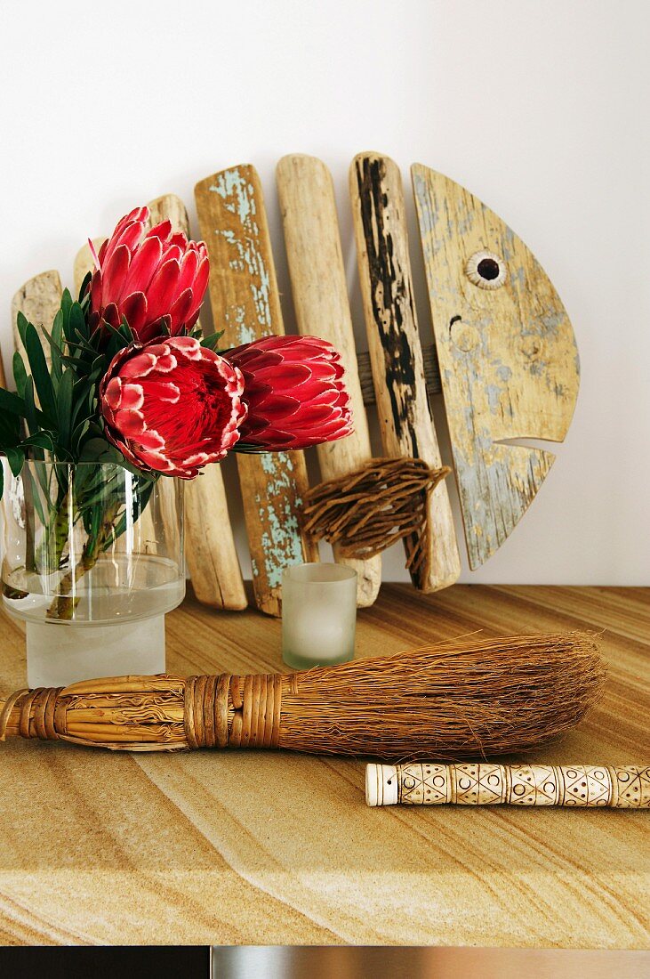 Protea Blumen in Vase und rustikaler Handbesen auf Tisch, im Hintergrund künstlerische Holzarbeit in Fischform an Wand gelehnt