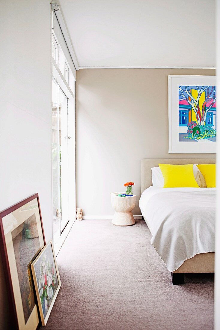 Double bed, splashes of colour provided by yellow scatter cushions and framed pictures leaning against wall in modern bedroom painted pale grey