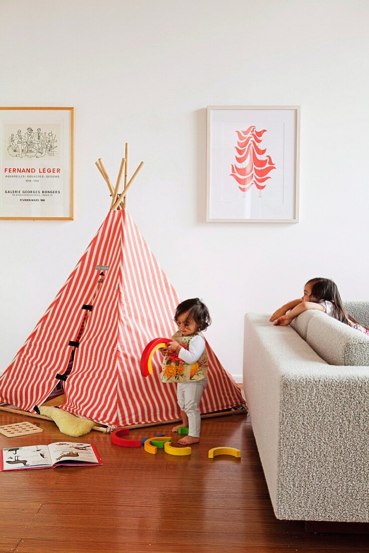 Kinder beim Spielen vor aufgebautem Indianerzelt mit rotweiss gestreiftem Stoff auf Parkettboden im Wohnzimmer