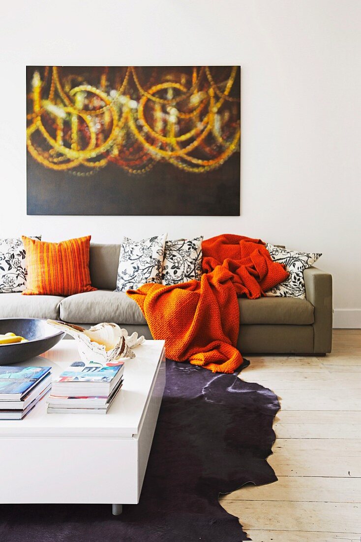 Low white coffee table on animal-skin rug, orange blanket and retro-style scatter cushions on sofa below modern painting on white wall