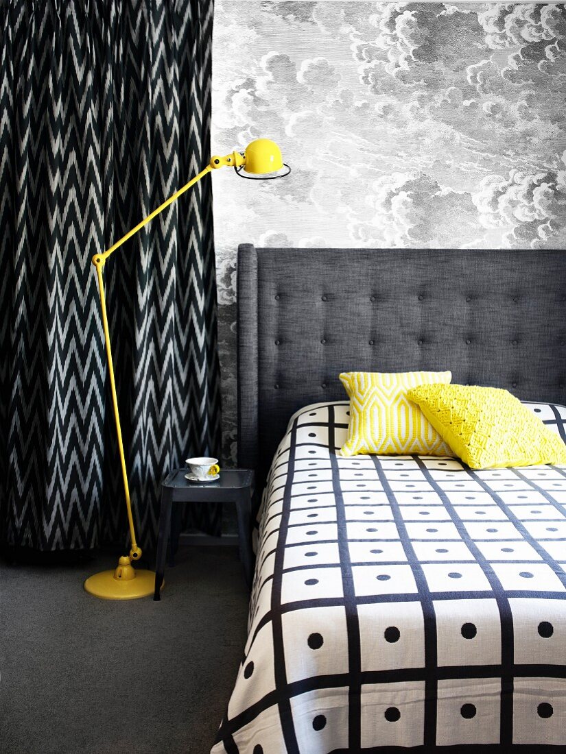 Double bed with grey upholstered headboard next to yellow, retro standard lamp against wall with cloud-patterned wallpaper