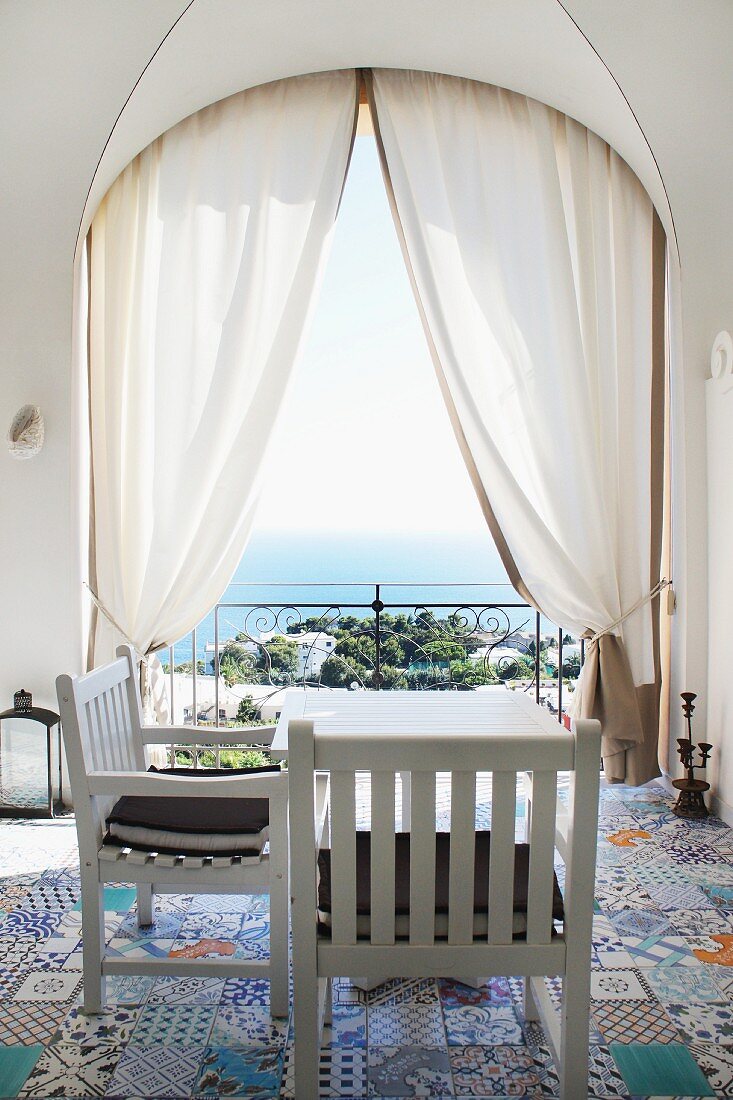 weiße Holzstühle mit Tisch auf bunt gemusterter, alter Bodenfliesen in Loggia mit Blick auf den Golf von Capri