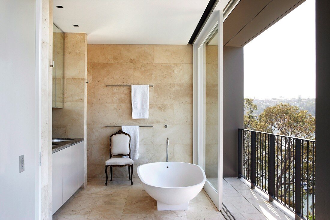 Freistehende Badewanne neben offener Balkonschiebetür in modernem Bad mit sandfarbenen Fliesen an Wand und Boden