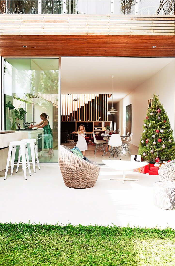 Weihnachtsbaum auf sonniger Terrasse vor offener Glas Schiebetür und Blick in Wohnraum auf Familie