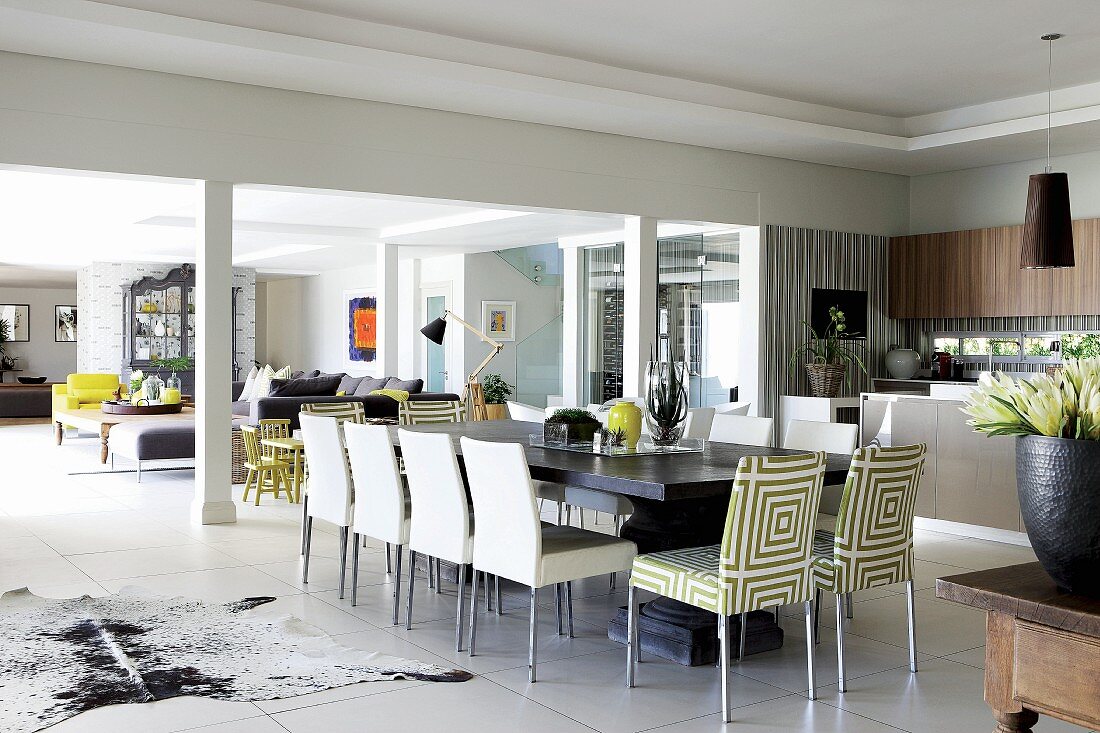 Langer Esstisch mit Polsterstühlen vor eleganter Küche; im Hintergrund Sitzbereich mit Sofalandschaft im grossen, offenen Wohnraum