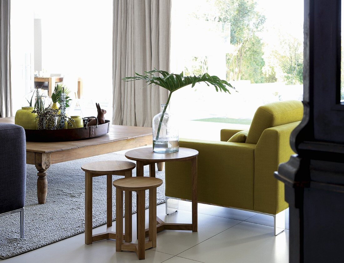 Beistelltische aus hellem Holz neben einem gelbgrünem Sessel und Tablett mit Vasen und Accessoires auf breitem Couchtisch