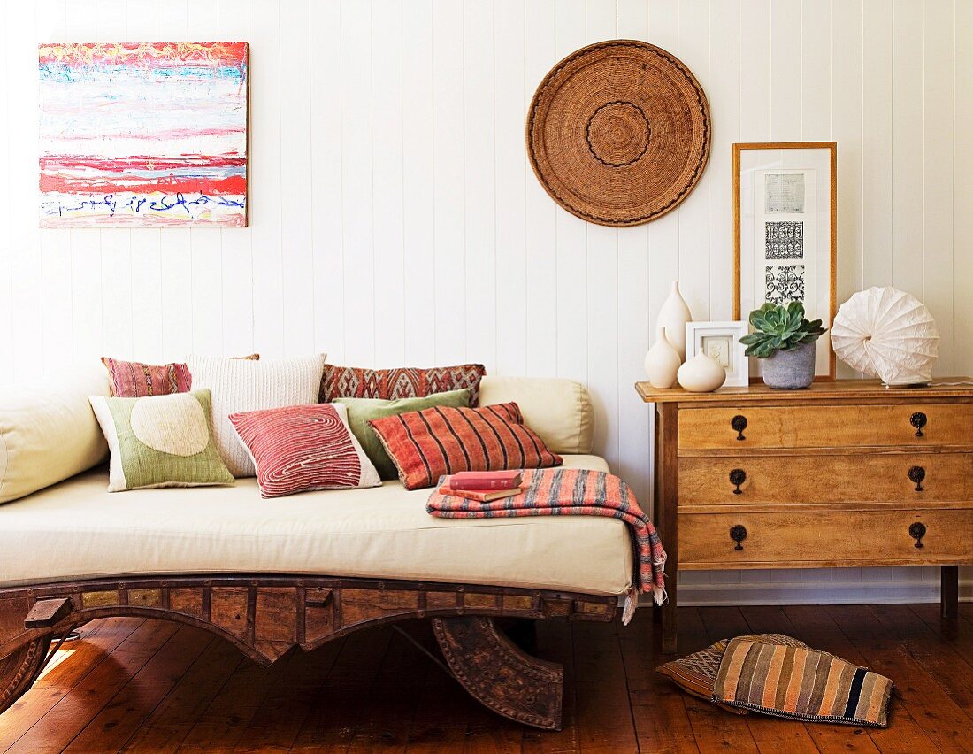 Daybed with pale cushions and colourful scatter cushions on wooden frame next to chest of drawers against white, wooden wall