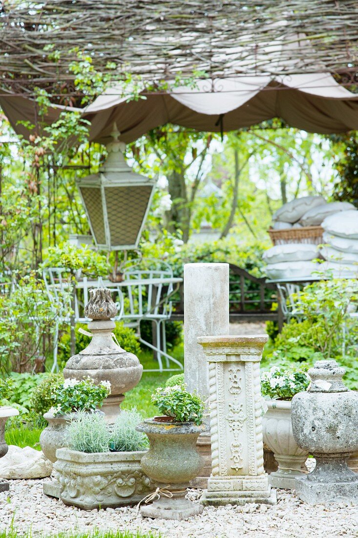 Zum Verkauf angebotene Steintöpfe und Säulen im Antikstil auf einer Kiesfläche im Garten; Laterne und weiße Metallstühle im Hintergrund