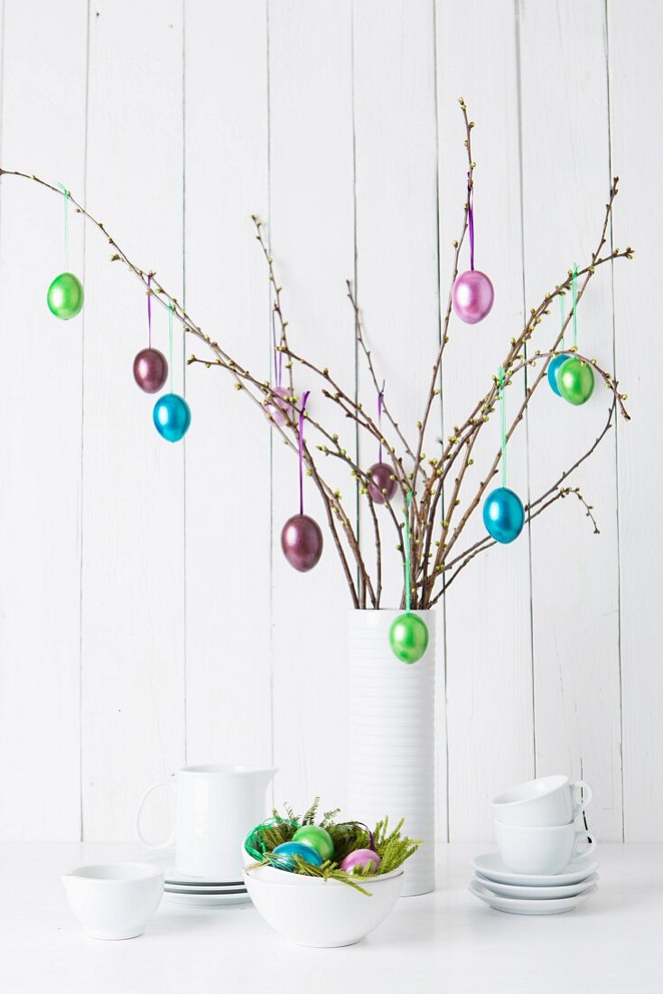 Glänzende Ostereier an Zweigen aufgehängt in weisser Vase und Geschirr auf Ablage vor weisser Holzwand