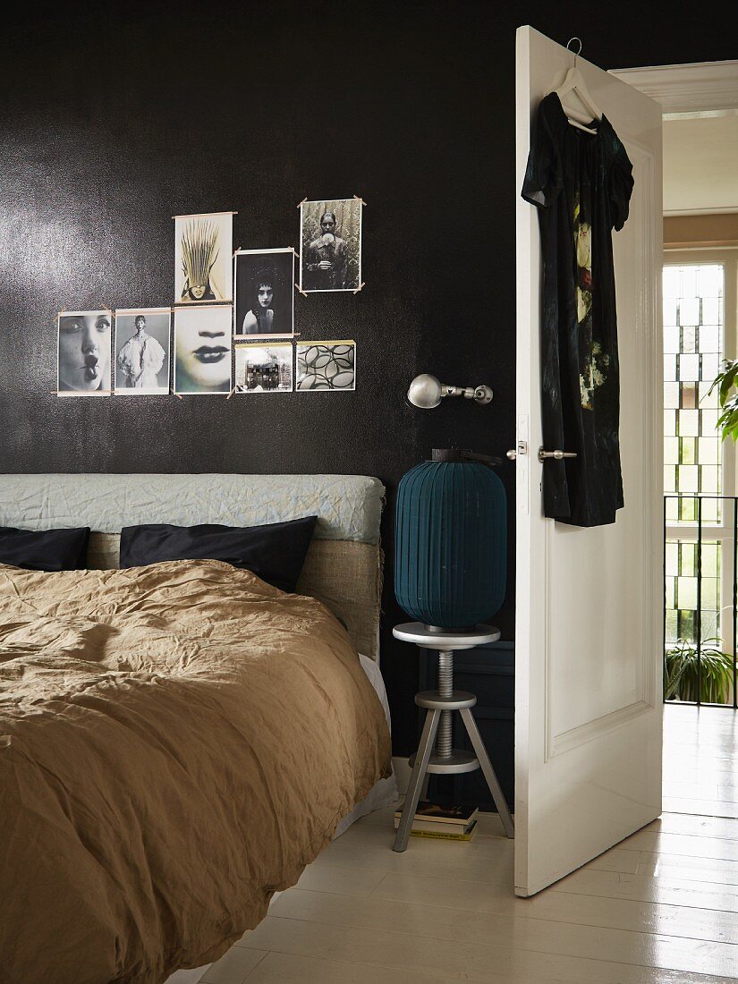 Bett mit Kopfpolster vor schwarz getönter Wand mit angehefteten Fotos, daneben die offene Zimmertür