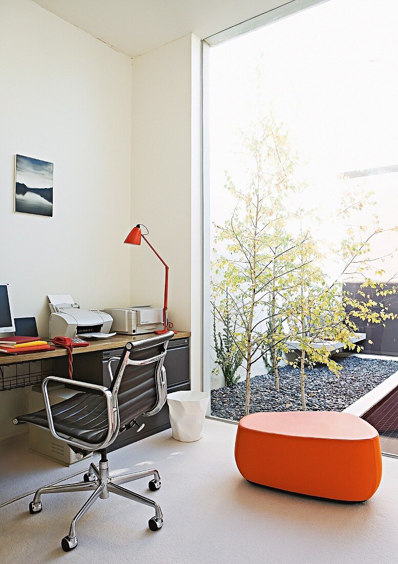 Classic office chair with black leather seat and orange, designer pouffe in home office; glass wall facing courtyard with birch trees in gravel bed