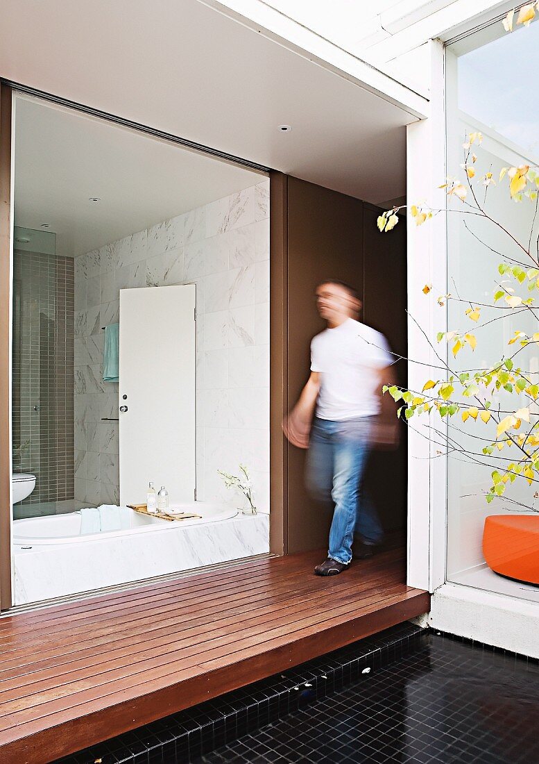 Blick von Innenhof mit Holzpodest durch rahmenlose Verglasungen in weisses Bad und gegenüberliegendes Homeoffice; mittig unscharf ein Mann in Bewegung