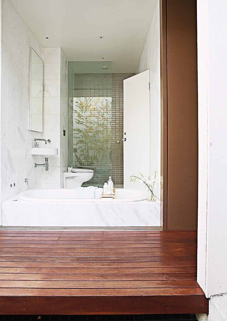 View from courtyard with wooden platform through frameless glass wall into bathroom