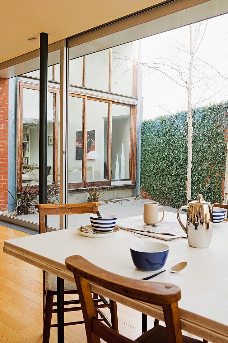 Mit modernem, blauweissem Geschirr für das Frühstück gedeckter Esstisch und Holzstühle vor Glasfassade mit Blick auf Innenhof und Übereckfassade