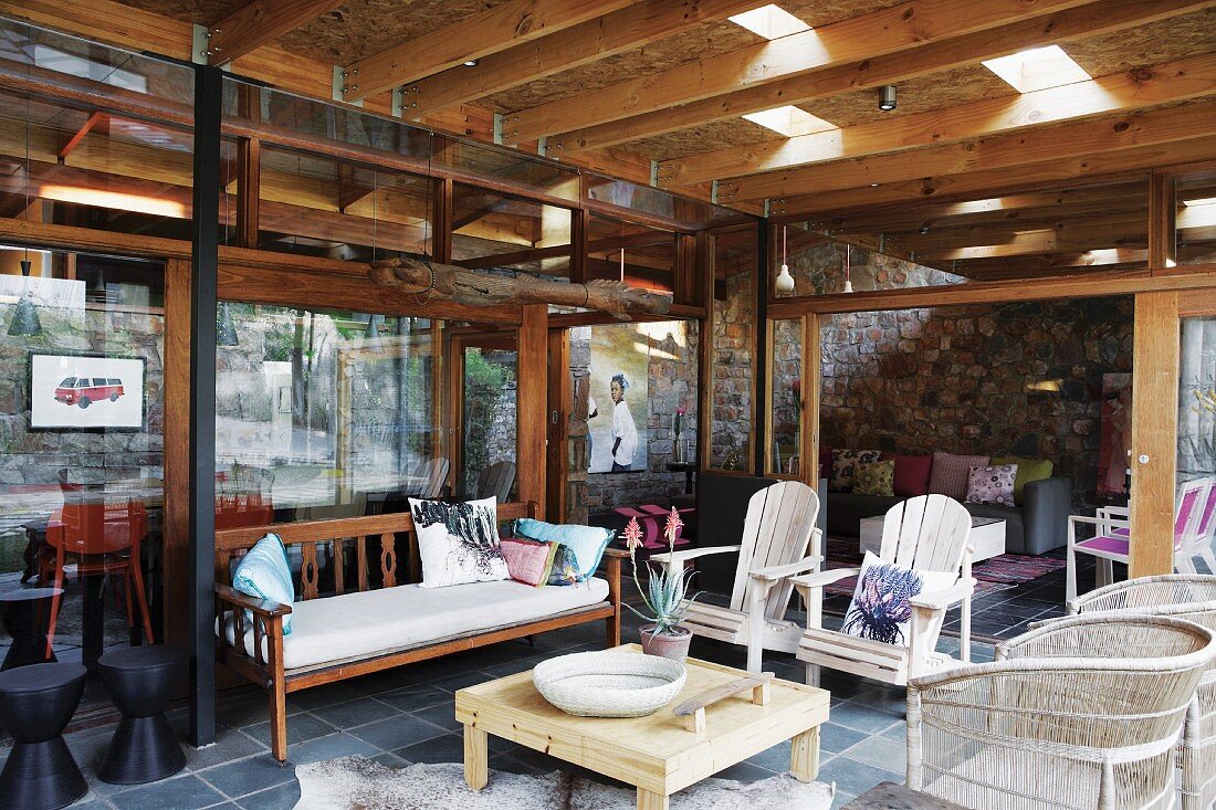 Mit Holzbank, Armlehnstühlen und Geflechtsesseln möblierte Terrasse vor der Glasfassade eines Wohnhauses; Holzdach mit Lichtöffnungen