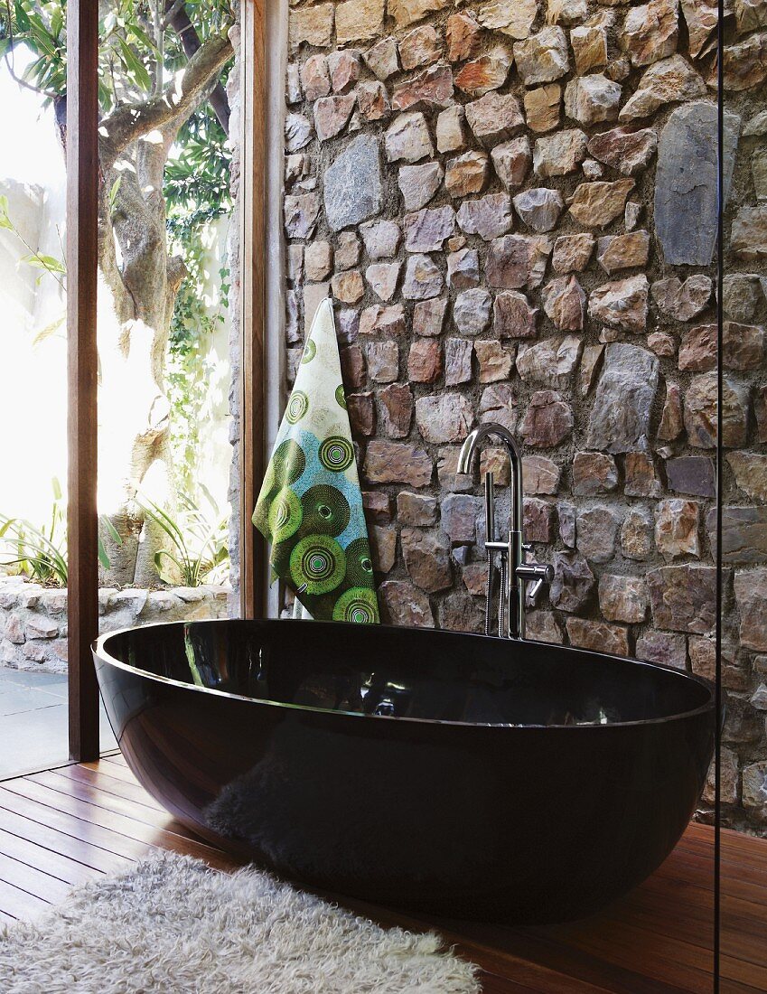 Designer bathroom with black, free-standing tub against stone wall and glass wall with view of garden terrace