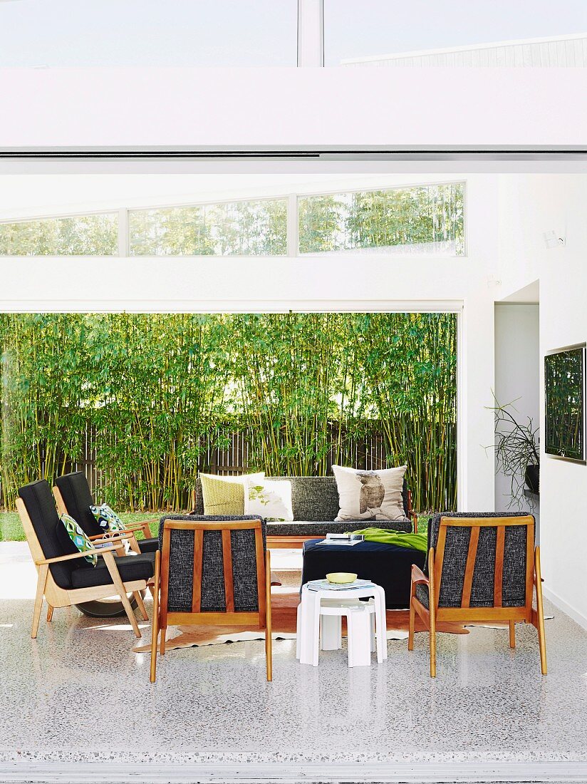 60s armchairs and sofa on terrazzo floor in bright interior; view of bamboo hedge and screen through wide, sliding glass wall