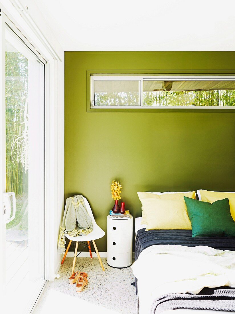 Cylindrical chest of drawers and classic chair for hanging clothes next to double bed; transom window with view of bamboo hedge in green-pained wall