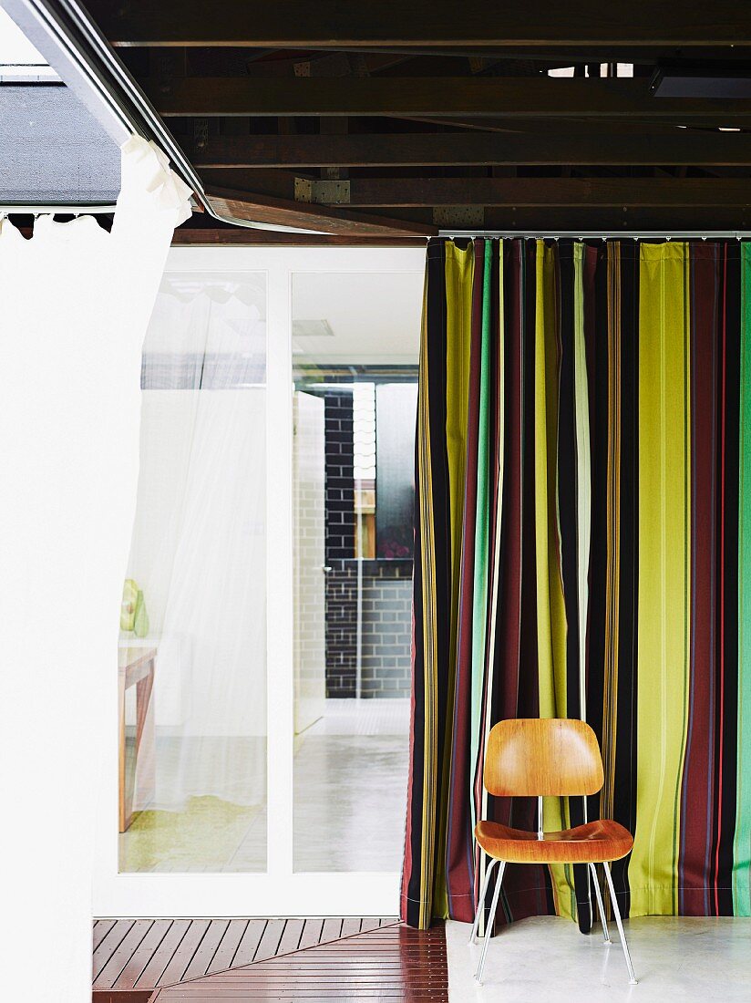 Übereck liegende Räume mit umlaufenden Holzstegen und Moskitovorhängen an einem Innenhof; Klassikerstuhl (LCM) von Eames vor gestreiftem Sichtschutzvorhang
