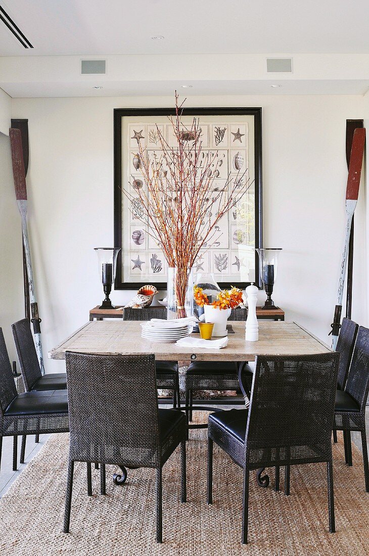 Oars and pictures of shells and starfish in large frame as maritime decor behind large dining table with stone top and dark chairs with leather seats