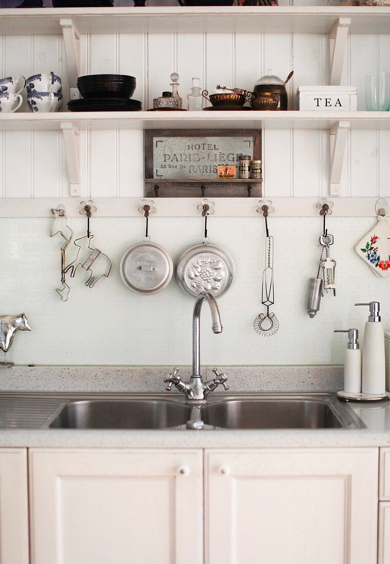 Doppelspülbecken mit weißem Unterschrank vor Hakenleiste mit Küchenutensilien unter Holzbord an Wand