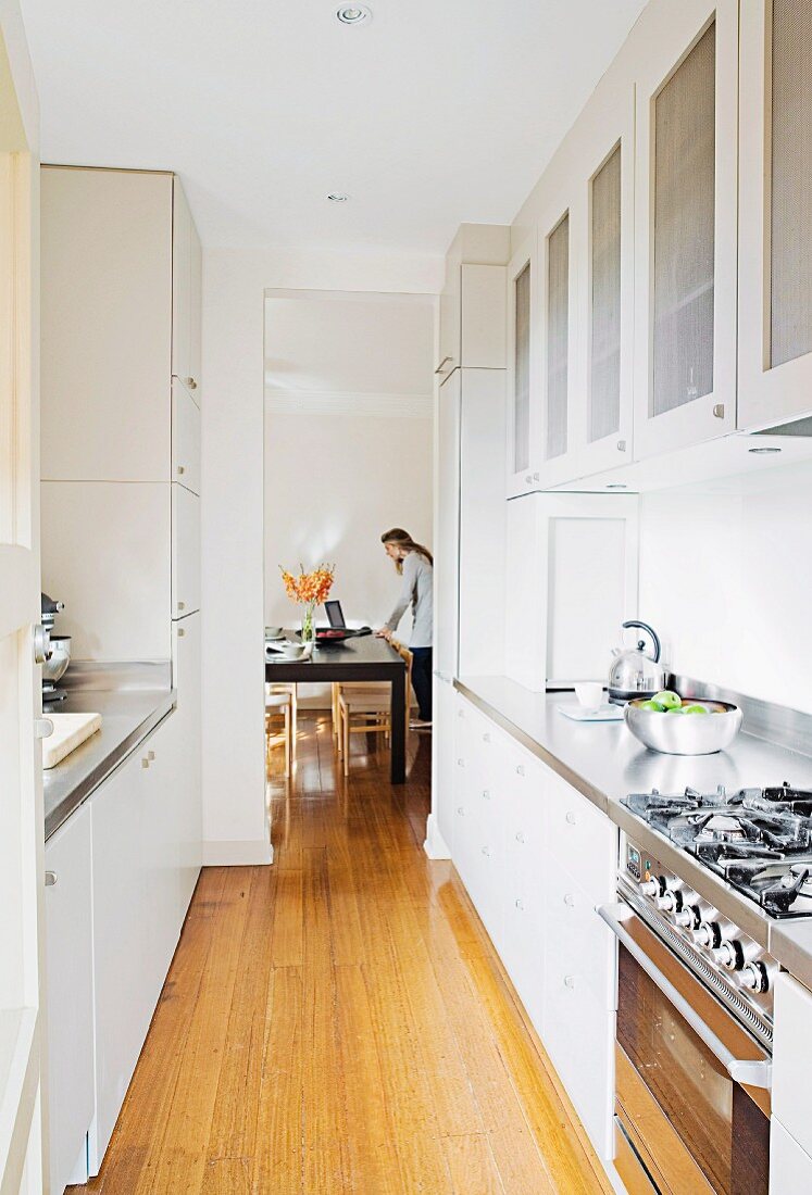 Narrow, white, fitted kitchen with continuous wooden floor leading into dining room; woman next to table in background