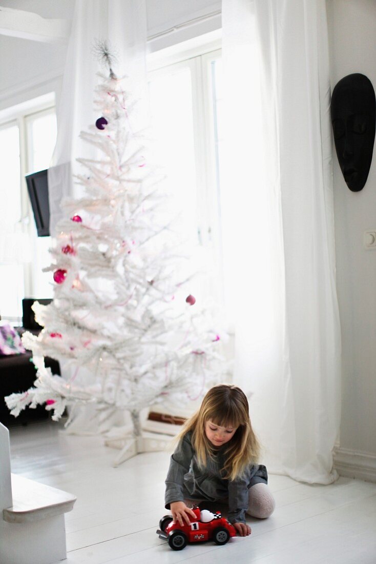 Mädchen mit Spielzeugauto auf weißem Dielenboden, dahinter weisser, künstlicher Weihnachtsbaum mit violetten Kugeln