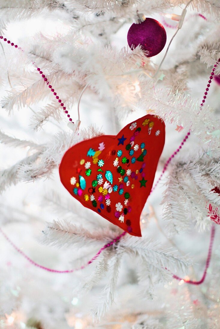 Rotes gebasteltes Herz mit bunten Pailletten und violette Kugel an weißem, künstlichem Weihnachtsbaum