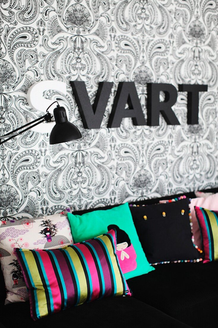 Scatter cushions with various patterns on black sofa below ornamental letters on patterned wallpaper