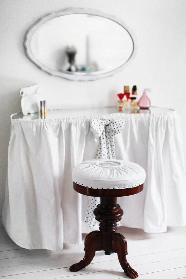 Antique piano stool with white seat cushion at curved dressing table with white curtain