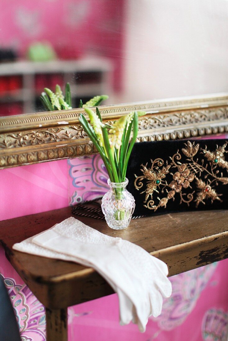 White lady's gloves and small, crystal vase of spring flowers on console table below mirror