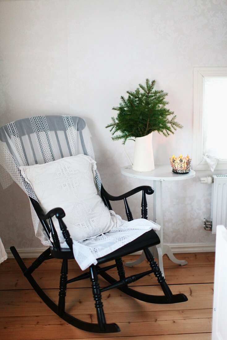Black-painted wooden rocking chair with white cushion next to water jug of fir branches on side table