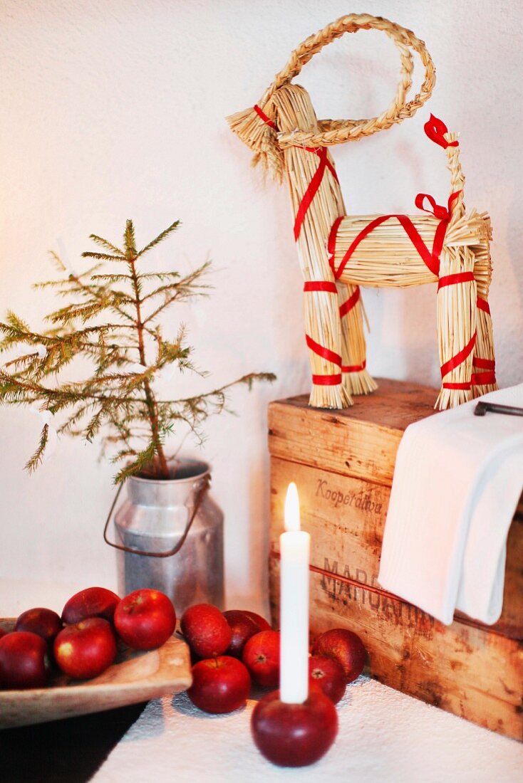 Festive arrangement of red apples and candle stuck in apple in front of straw reindeer decorated with red ribbons on wooden trunk in corner