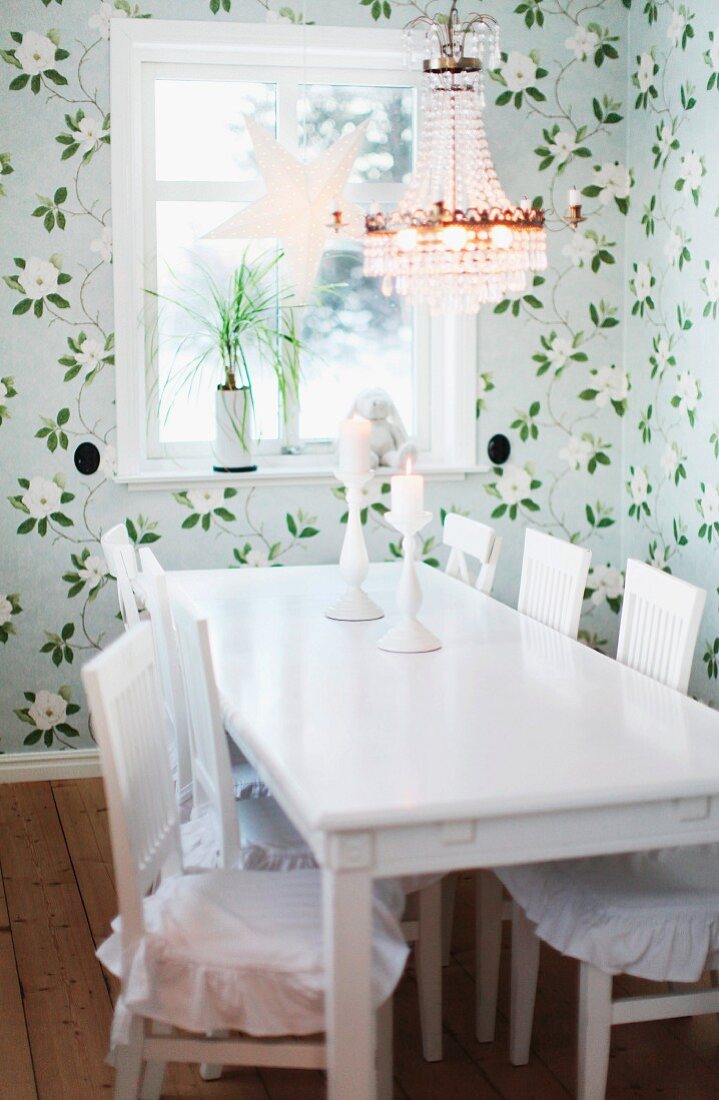 Esstisch und Stühle in weiss lackiert, darüber traditioneller Kronleuchter mit Glasschmuck in ländlicher Esszimmerecke mit pastelliger Blumentapete, am Fenster weihnachtlicher Dekostern