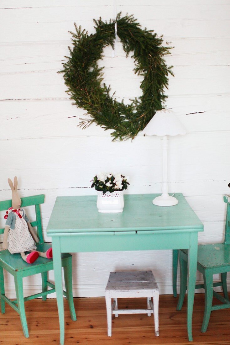 Wooden table and chairs painted pastel turquoise below wreath of fir branches on white wooden wall